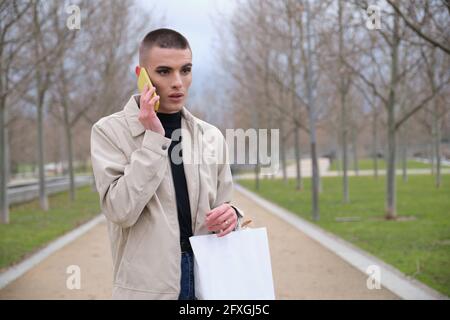 Jeune homme portant un maquillage tenant des sacs de shopping et parlant sur un smartphone dans un parc. Queer personne. Banque D'Images