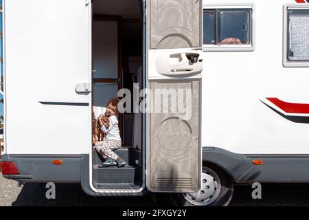 petit garçon aux cheveux bouclés souriant assis dans la porte de la maison de moteur, tenant tendement son animal de compagnie. Banque D'Images