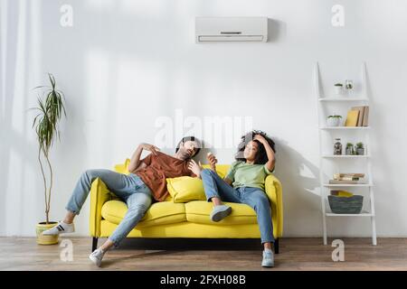 un couple multiethnique en sueur assis sur un canapé jaune et souffrant de chaleur en été Banque D'Images