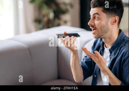 Jeune sympathique sympathique cacasien gars dans des vêtements décontractés assis sur le canapé et l'enregistrement d'un message audio ou la conversation téléphone intelligent sur haut-parleur avec un ami ou une famille tout en faisant des gestes avec sa main Banque D'Images