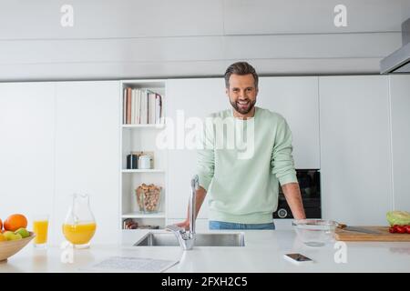 homme joyeux souriant à l'appareil photo près du jus d'orange et du lavabo dans la cuisine Banque D'Images