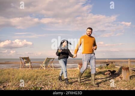 Couple heureux avec le vin tenant les mains marchant sur la plage ensoleillée Banque D'Images