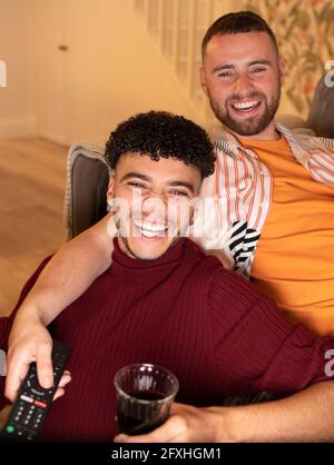 Un couple de hommes gais heureux regardant la télévision et buvant du vin rouge à la maison Banque D'Images