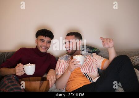 Un couple de hommes gay heureux qui boit du chocolat chaud en regardant la télévision sur le canapé Banque D'Images