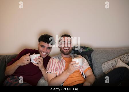 Un couple de hommes gay heureux qui boit du chocolat chaud et qui regarde la télévision sur le canapé Banque D'Images