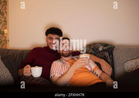 Couple gay affectueux et heureux avec chocolat chaud regardant la télévision sur le canapé Banque D'Images