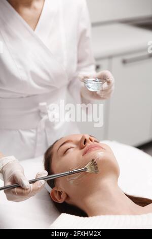 L'esthéticienne brosse le gel avec la brosse sur le patient de la clinique de beauté, dermatologue utilisant la brosse pour insérer le produit cosmétique sur le visage du client, préparateur pour la peau Banque D'Images