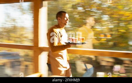 Un barista masculin porte un plateau de café le long de la fenêtre ensoleillée du café d'automne Banque D'Images