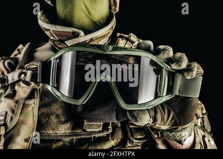 Photo d'un soldat dans des munitions blindées de niveau 3, gants tactiques tenant des lunettes tactiques sur fond noir. Banque D'Images