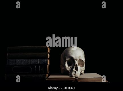 Photo du crâne humain de sorcellerie sur des livres anciens et des livres empilés sur fond noir. Banque D'Images