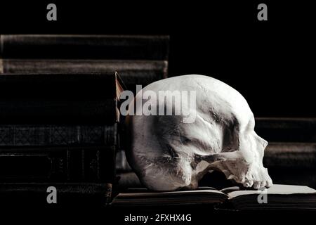 Photo du crâne humain de sorcellerie sur des livres anciens et des livres empilés sur fond noir. Banque D'Images