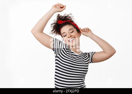 Une jeune femme insouciante, vêtue d'une tenue de pinup, dansant et levant les mains détendues, profitant de la musique de fête, s'amusant, debout sur fond blanc Banque D'Images