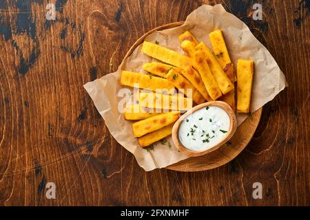 Croustilles de polenta faites maison frites avec sel de mer, parmesan, thym, romarin et sauce au yaourt. Polenta typiquement italienne. Bâtonnets de maïs frits. Dos en bois Banque D'Images