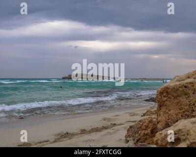 Les personnes qui nagent sur les vagues de la mer lors d'une journée couverte. Plage de Nissi pittoresque avec de l'eau cyan troublée, une petite île rocheuse et un ciel sombre et nuageux à Ayia Napa Banque D'Images