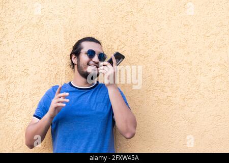 jeune latino barbu avec des lunettes de soleil appuyé contre un mur jaune envoyant un message vocal. air frais Banque D'Images