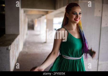 Bonne fille dans une robe verte lumineuse avec un maquillage de gritter coloré et des tresses africaines arc-en-ciel. Milo sourit et tourne dans les rues d'une grande ville plus chaude Banque D'Images