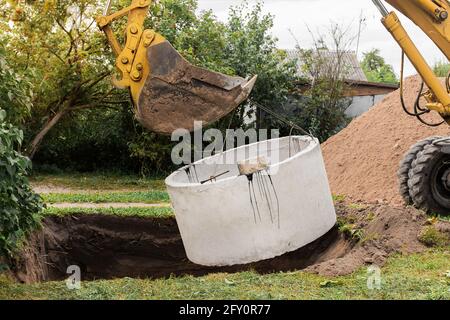 Pelle hydraulique avec un godet, s'abaissant dans la fosse sur des câbles en acier anneau d'égout en béton. Construction ou réparation d'un égout. Banque D'Images