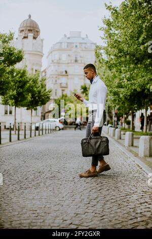 Beau jeune homme d'affaires afro-américain utilisant un téléphone portable pendant céossier une rue Banque D'Images