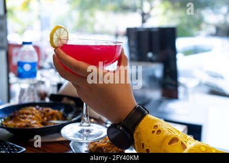 Jeune femme indienne en robe jaune montre tenant un grand verre de vin rempli d'un cocktail rouge de cerise de fraise avec une tranche de citron Banque D'Images