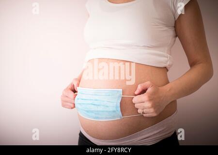 Femme enceinte mettant un masque bleu sur le ventre pendant la pandémie Covid-19. Coronavirus en question de grossesse, symbole de protection. Arrière-plan de la vue latérale Banque D'Images