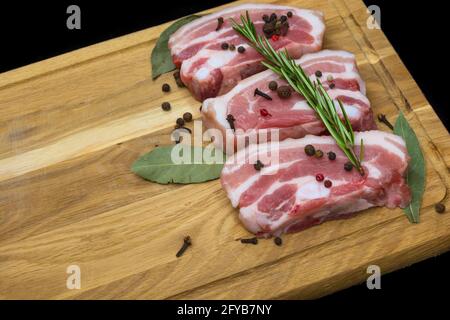 Poitrine de porc crue, hachée sur un plan de cuisine, parsemée d'épices, de clous de girofle et de feuilles de Laurier. Arrière-plan noir. Banque D'Images