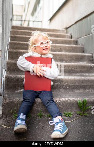 fille en tricot chaud blanc veste et baskets tient un grand livre. enfant porte des lunettes près de l'école. Jardin d'enfants, concept de l'école élémentaire. Banque D'Images