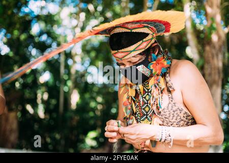 Indien de la tribu Pataxó avec une coiffe en plumes et un masque protecteur contre la pandémie de Covid-19. Femme indigène du Brésil Making Crafts Banque D'Images