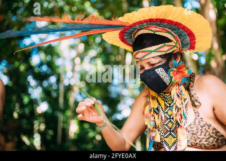 Indien de la tribu Pataxó avec une coiffe en plumes et un masque protecteur contre la pandémie de Covid-19. Femme indigène du Brésil Making Crafts Banque D'Images