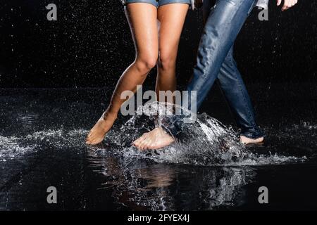 vue partielle des jambes pieds nus hommes et femmes mouvements dans la flaque avec éclaboussures d'eau sur fond noir Banque D'Images