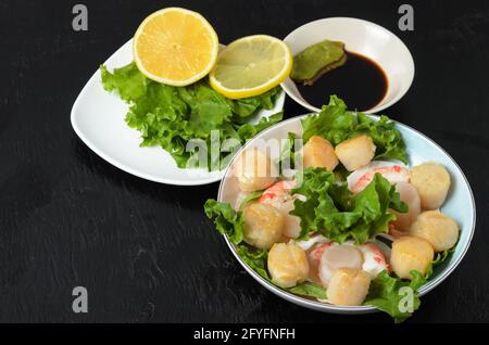 Viande de pétoncle frite aux crevettes bouillies dans une assiette en céramique avec laitue, citron, sauce soja et wasabi sur fond sombre. Mise au point sélective. Banque D'Images