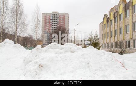 Une grande dérive des neiges s'est accumulée sur une rue de la ville avec des immeubles en hauteur. Un bloc de neige avec des morceaux sales se trouve dans la ville dans la cour d'un immeuble d'appartements. Jour d'hiver sombre. Banque D'Images