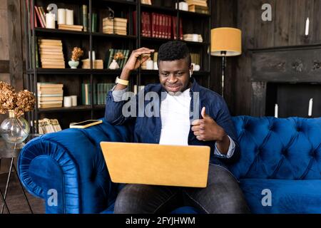 Jeune homme d'affaires élégant assis sur un canapé, montrant le pouce jusqu'à la vidéoconférence en ligne avec un ordinateur portable jaune pour des collègues. Indépendant ou Banque D'Images