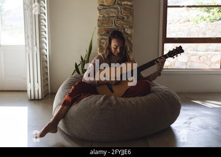 Bonne femme caucasienne assise sur un beanbag jouant de la guitare acoustique séjour cottage ensoleillé Banque D'Images