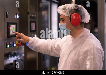 CAPUA, ITALIE - 23 mai 2021: Employé travailleur homme en casaque masqué et de protection panneau de commande de la machine et des commandes. Capua, Italie, mai Banque D'Images