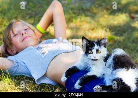 Flou blonde petite fille couché sur l'herbe avec chat, noir et blanc petit chatte mignon avec les yeux jaunes. Nature vert fond d'été. Fille caresse pe Banque D'Images