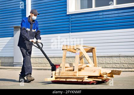 Un homme en vêtements de travail et un masque médical porte des planches en bois sur une charrette sur le territoire d'une base industrielle pendant une journée d'été. Travailler conformément aux normes sanitaires Banque D'Images