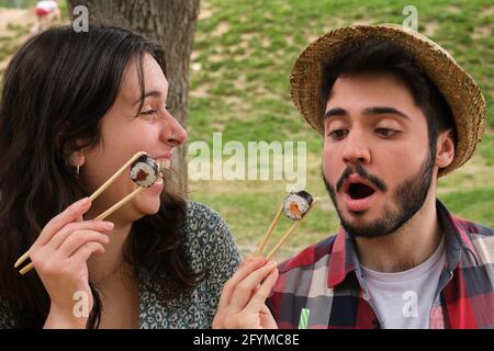 Un jeune couple heureux riant et mangeant des sushis dans un parc pique-nique par une belle journée d'été. Banque D'Images