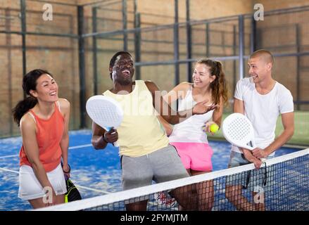 Heureux rire des hommes et des femmes de différentes nationalités dans les vêtements de sport avec des raquettes et des balles dans les mains parler amical près du filet sur le terrain de padel intérieur Banque D'Images