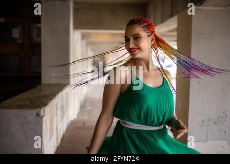 Bonne fille dans une robe verte lumineuse avec un maquillage de gritter coloré et des tresses africaines arc-en-ciel. Milo sourit et tourne dans les rues d'une grande ville plus chaude Banque D'Images