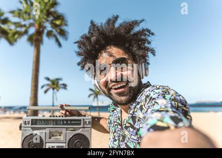 Jeune latino s'amusant à prendre un selfie avec un smartphone mobile tout en écoutant de la musique avec un casque et une boombox sur la plage pendant les vacances d'été Banque D'Images