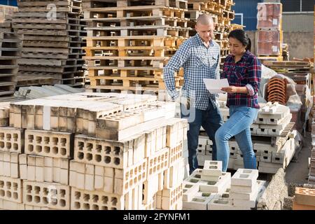 Femme préparatrice de commande avec des papiers collectant des produits dans l'entrepôt de matériaux de construction, homme travailleur l'aidant Banque D'Images