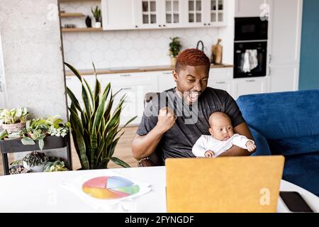 Travailleur indépendant avec bébé fils entre les mains ayant la vidéoconférence, entendre de bonnes nouvelles sur le projet, profiter des résultats du travail. Joyeux Banque D'Images