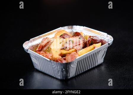 Conteneur de nourriture pour la livraison de nourriture du restaurant. Pommes de terre frites avec bacon fumé et saucisses dans un récipient en aluminium sur fond noir. Banque D'Images