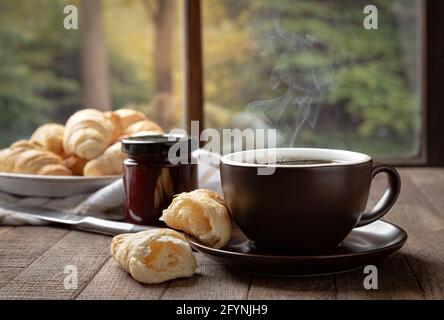 Tasse de café chaud et croissants sur une table en bois par une fenêtre avec une scène rurale d'arrière-plan Banque D'Images