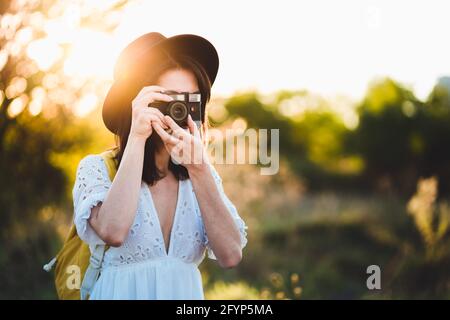 Hipster jeune femme touriste utilisant un appareil photo rétro vintage. Jeune femme à la mode avec sac à dos touristique prenant des photos dans la nature au coucher du soleil. M Banque D'Images