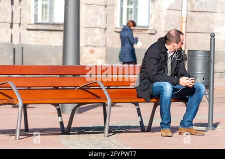 Timisoara, Roumanie - 14 mars 2016 : homme assis sur un banc dans la rue et écoutant de la musique depuis son téléphone. De vraies personnes. Banque D'Images