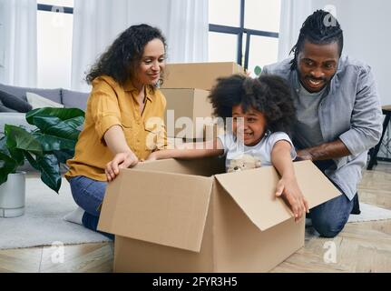 Adorable petite fille afro-américaine assise dans une boîte en carton, jouant avec son parent tout en se déplaçant dans une nouvelle maison Banque D'Images