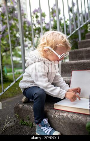 Retour à l'école. Une petite fille intelligente lit un grand livre. L'enfant s'assoit sur les marches de l'école avec des lunettes. Manuel ouvert devant l'enfant. Banque D'Images