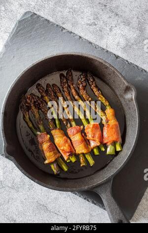 Asperges enrobées de bacon cuit dans une poêle en fonte. Sur une planche à découper en ardoise Banque D'Images