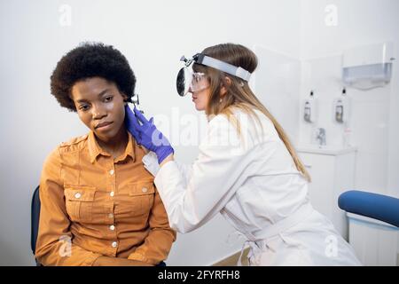 Otite, concept de traitement de la perte auditive. Jeune femme confiante otorhinolaryngologiste examinant l'oreille douloureuse de sa patiente afro-américaine avec otoscope dans une clinique moderne Banque D'Images